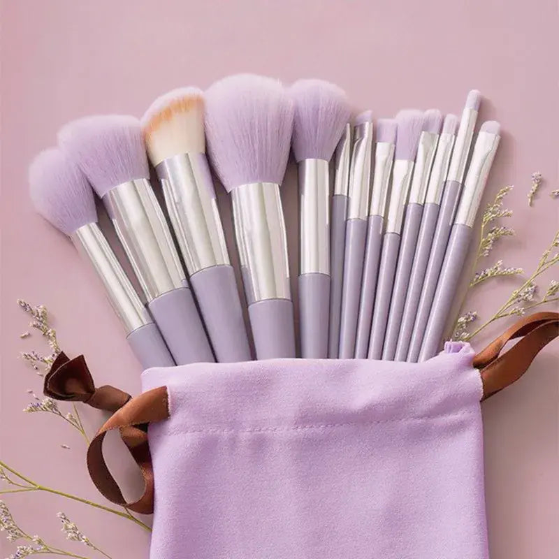 A set of makeup brushes with pale lavender bristles and silver handles, arranged in a matching lavender fabric pouch with a brown ribbon, on a soft pink background.