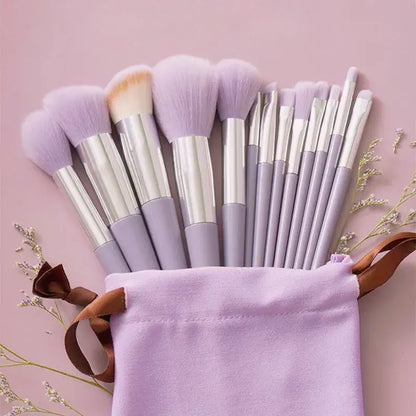 A set of makeup brushes with pale lavender bristles and silver handles, arranged in a matching lavender fabric pouch with a brown ribbon, on a soft pink background.
